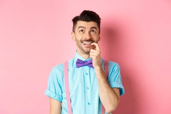 Образ усміхненого чоловіка, який робить вибір, виглядає мрійливим і щасливим, кусаючи нігті з спокусливим виразом, бажаючи чогось, стоїть над рожевим тлом — стокове фото