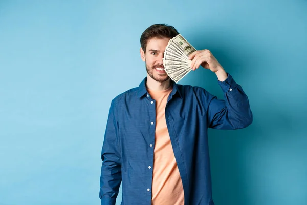Happy young guy covering half of face with dollars and smiling, winning prize money, standing on blue background