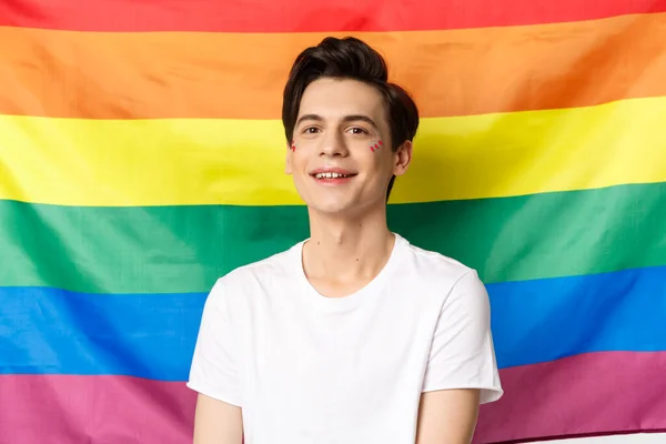 Hermoso hombre gay con brillo en la cara, sonriendo feliz y orgulloso a la cámara, de pie contra la bandera del orgullo del arco iris, lgbtq derechos de la comunidad y el concepto de la gente —  Fotos de Stock