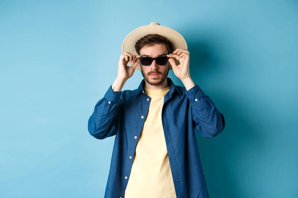 夏休みにサングラスをかけて青い背景に立つわら帽子のハンサムな観光客 — ストック写真