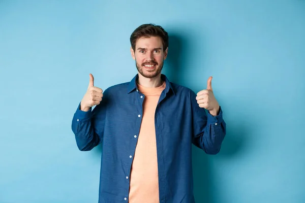 Knappe glimlachende man die ondersteunend, duimen omhoog in goedkeuring, lof goed werk, staande gelukkig op blauwe achtergrond — Stockfoto