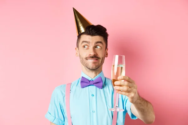 Vakantie en feest concept. Vrolijke jongeman viert verjaardag in feesthoed, denken aan spraak, glas chamapgne heffen voor toast, roze achtergrond — Stockfoto