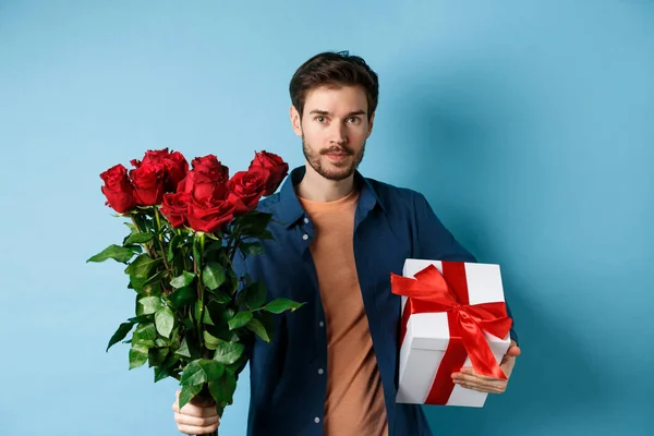 Romance y San Valentín. Hombre presentando ramo de rosas rojas a amante. Novio traer flores y regalo en fecha romántica, de pie sobre fondo azul —  Fotos de Stock