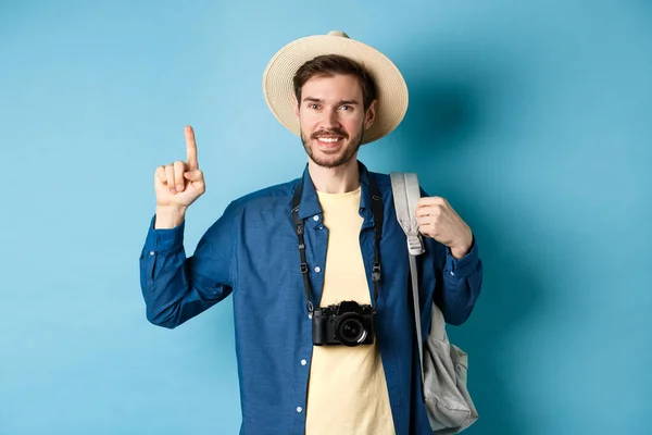 Guapo turista feliz en sombrero de verano, la celebración de la mochila y la cámara, señalando el dedo hacia arriba en el logotipo, recomendando agencia de viajes o lugar de vacaciones, fondo azul — Foto de Stock