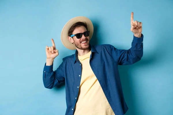 Divertido turista bailando de vacaciones, señalando con los dedos hacia arriba, usando sombrero de verano y gafas de sol, fondo azul — Foto de Stock