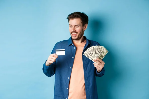 Hombre joven guapo mostrando dinero en efectivo y mirando a la tarjeta de crédito de plástico, prefieren el pago sin contacto, de pie sobre fondo azul — Foto de Stock