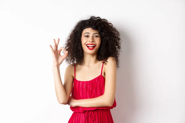 Χαρούμενη καυκάσια γυναίκα με σγουρά μαλλιά και μακιγιάζ, φορώντας κομψό κόκκινο φόρεμα, που δείχνει OK σημάδι και χαμογελώντας σε έγκριση, στέκεται πάνω από το λευκό φόντο — Φωτογραφία Αρχείου