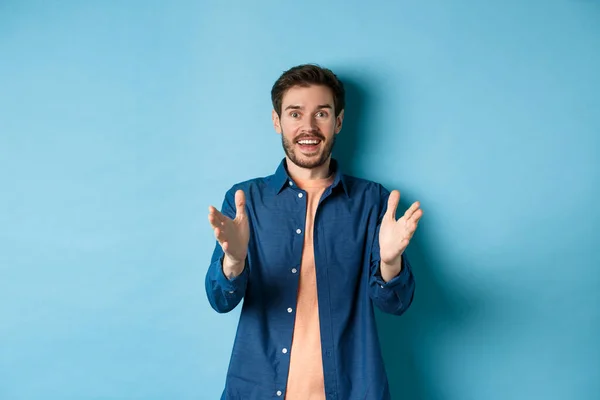 Vrolijke jongeman die zijn handen op de camera richt en glimlacht, blij voor je is, mooi werk prijst, op een blauwe achtergrond staat — Stockfoto