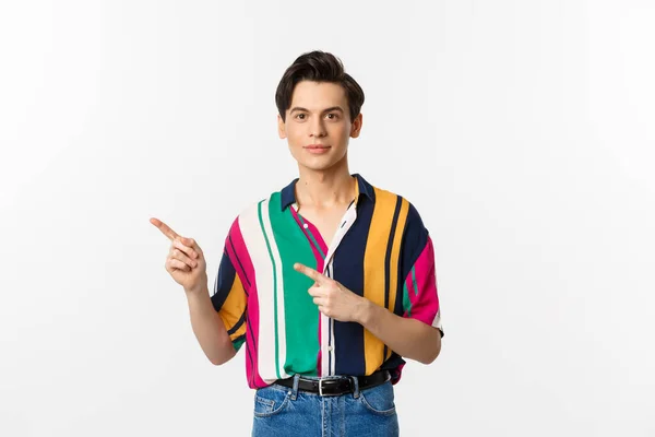 Kép elegáns androgén férfi mosolygós, ujjal balra mutató logó, bemutató promo ajánlat, áll a fehér háttér felett — Stock Fotó
