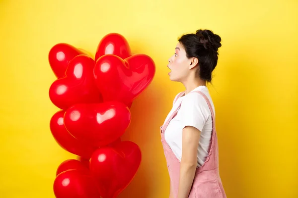 Sevgililer günü ve ilişki konsepti. Şaşırmış Asyalı kızın profili logoya bakıyor, kalp balonlarının yanında duruyor, sarı arka plan. — Stok fotoğraf