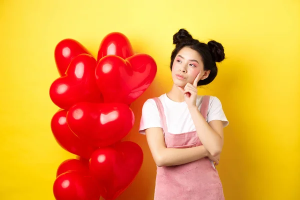 Valentines day concept. Pensive asian woman looking left, thinking about romantic date, standing near red hearts balloons on yellow background