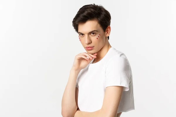 People, lgbtq and beauty concept. Beautiful androgynous male model with glitter on face, wearing crop top, looking with piercing eyes at camera, standing over white background — Stock Photo, Image