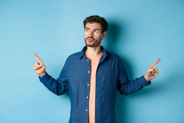 Uomo indeciso in abiti casual fare la scelta, puntando il dito lateralmente e guardando pensoso, in piedi su sfondo blu — Foto Stock