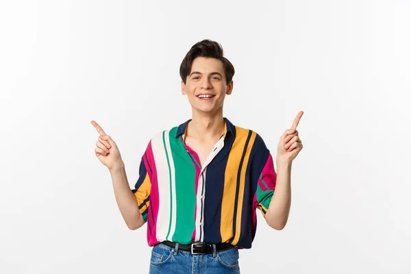 Happy young gay man pointing fingers sideways, showing two choices and smiling, standing against white background — Stock Photo, Image