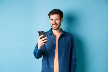 Çekici beyaz adam cep telefonu kullanıyor, mutlu görünüyor ve kameraya gülümsüyor, mavi arka planda duruyor.