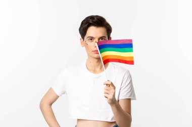 Lgbtq topluluğu. Yüzünde fiske olan çekici ibne, gururlu gökkuşağı bayrağı sallıyor ve kameraya bakıyor, beyaz arka plana karşı mahsul tepesinde duruyor.