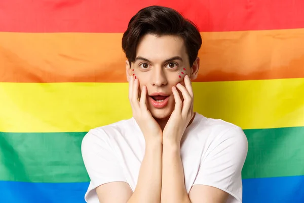 Close-up de belo homem gay morena em t-shirt branca, com maquiagem brilho, olhando para a câmera, tocando o rosto, de pé contra a bandeira do orgulho do arco-íris — Fotografia de Stock