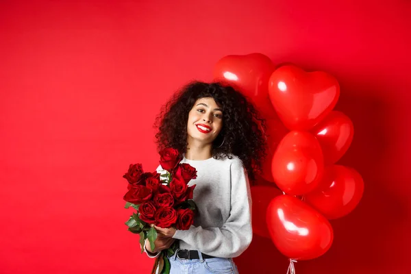 Holidays and celebration. Happy beautiful woman with curly hair, receive bouquet of roses and smiling, standing near party balloons, red background