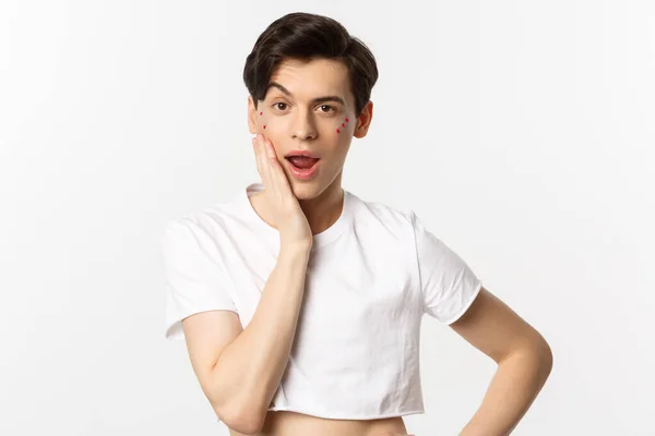 People, lgbtq and beauty concept. Sassy queer person with glitter on face looking cheeky at camera, touching face with hand, standing over white background — Stock Photo, Image