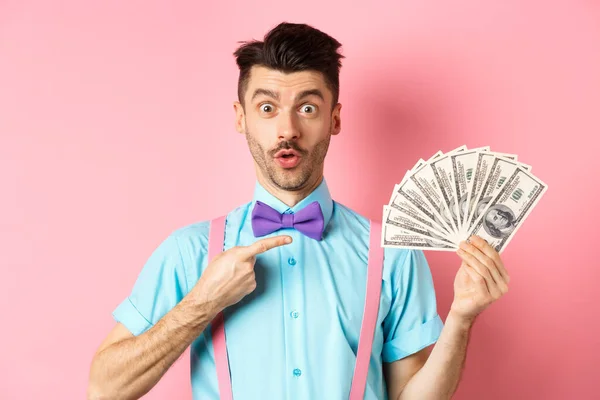 Hombre sorprendido mostrando gran premio de dinero, apuntando a los dólares y decir wow, mirar fijamente impresionado a la cámara, de pie sobre el fondo rosa — Foto de Stock