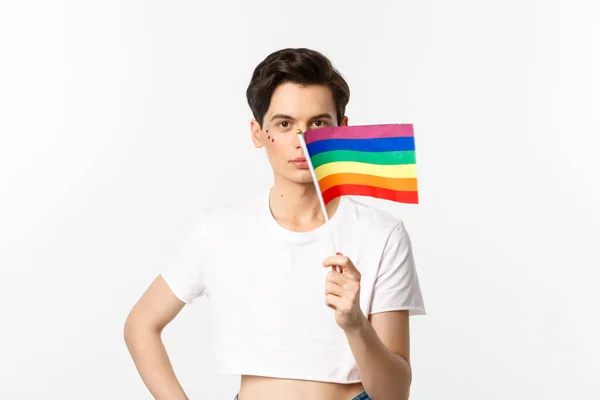 Lgbtq community. Attractive queer man with flitter on face, waving pride rainbow flag and looking at camera, standing in crop top against white background — Stock Photo, Image