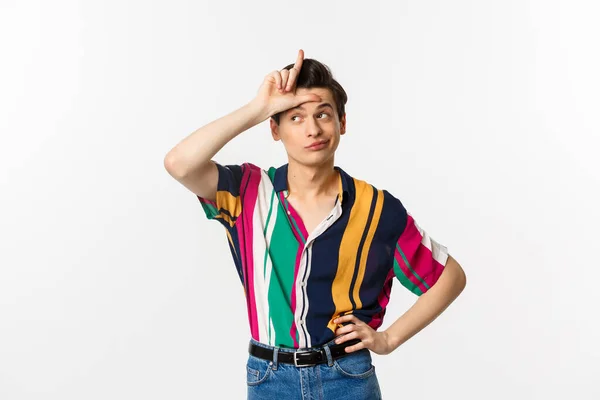 Image of arrogant young man showing loser sign on forehead to mock, looking away with pleased grin, standing over white background — Stock Photo, Image
