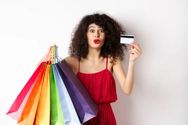 兴奋的女人，穿着红色衣服，拿着购物袋，展示塑料信用卡，购物打折，白色背景 — 图库照片