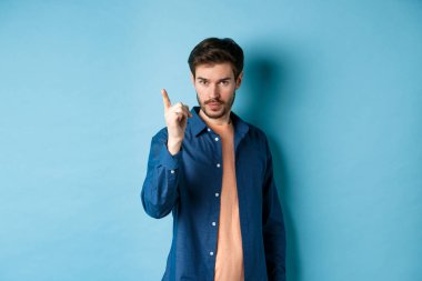 Ciddi bir genç adam azarlıyor, onaylamayan bir şekilde parmağını sallıyor, kötü bir şeyi uyarıyor veya yasaklıyor, mavi arka planda duruyor.
