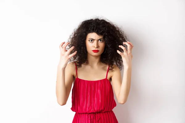 Θυμωμένη Ιταλίδα με σγουρά μαλλιά και κόκκινο φόρεμα, που βράζει από θυμό, κατσουφιάζει και σφίγγει τα χέρια εξοργισμένη, στέκεται σε λευκό φόντο — Φωτογραφία Αρχείου