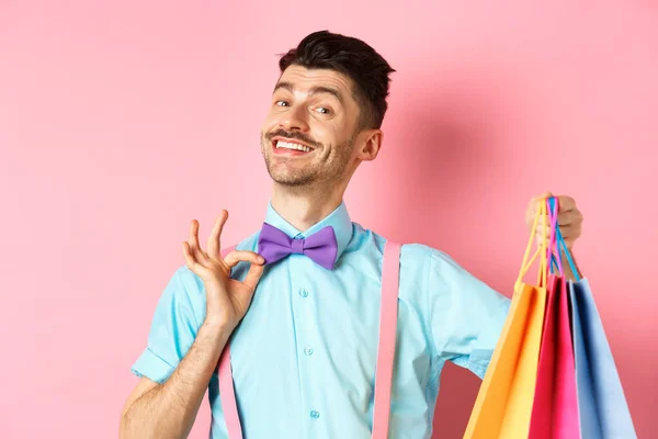 Chico sonriente confiado ajustando su pajarita y mostrando regalos en bolsas de compras, luciendo feliz, de pie sobre fondo rosa —  Fotos de Stock