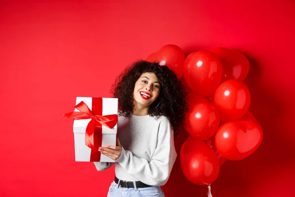 Vacanze e festeggiamenti. Buon compleanno ragazza in possesso di regalo e in posa vicino partito palloncini di elio, sorridente eccitato alla fotocamera, sfondo rosso — Foto Stock