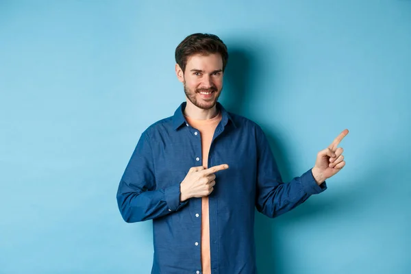 Glimlachende zelfverzekerde man met logo, wijzende vingers naar lege ruimte en kijkend naar de camera, staand op een blauwe achtergrond — Stockfoto