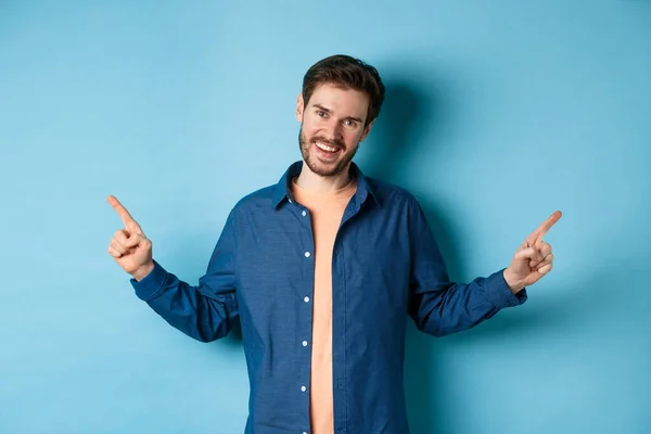 Knappe blanke man met baard, glimlachend met witte tanden en wijzende vingers zijwaarts, met twee keuzes, staand op een blauwe achtergrond — Stockfoto