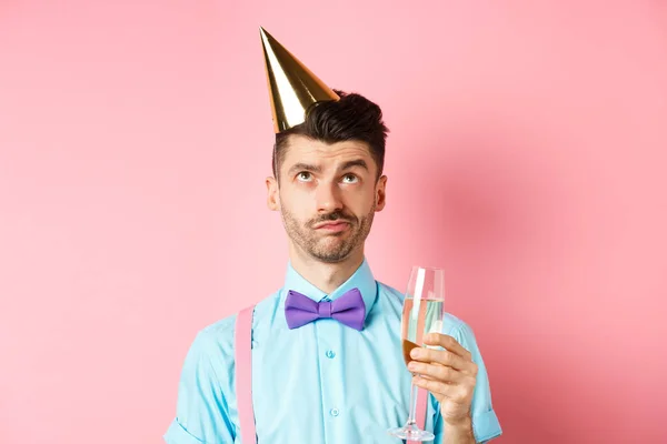 Vakantie en feest concept. Grumpy man met verjaardagsfeestmuts en een glas champagne, omhoog kijkend met sceptisch gezicht, staande op roze achtergrond — Stockfoto