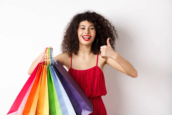 Μοντέρνα γυναίκα με κόκκινο φόρεμα πηγαίνει για ψώνια, κρατώντας τσάντες και δείχνει αντίχειρες επάνω, προτείνουμε κατάστημα, στέκεται σε λευκό φόντο — Φωτογραφία Αρχείου