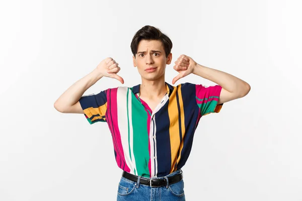 Disappointed queer gay judging something bad, showing thumbs down and grimacing displeased, dislike and disagree, standing over white background — Stock Photo, Image