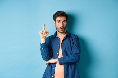 Yakışıklı genç adamın bir fikri var, plan yapıyor, parmağını kaldırıyor ve öneri diyor, mavi arka planda duruyor.