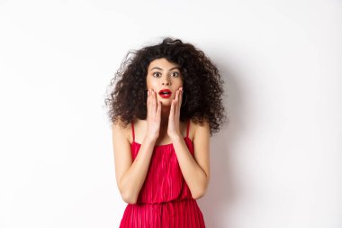 Heyecanlı kıvırcık saçlı kadın, inançsızlıkla yüz yüze, kameraya şaşkın şaşkın bakıyor, tanıtıma bakıyor, beyaz arka planda kırmızı bir kadın gibi duruyor.