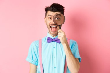 Papyonlu komik genç adam büyüteçten bakıyor ve dilini gösteriyor, mutlu gülümsüyor, pembe arka planda eğleniyor.