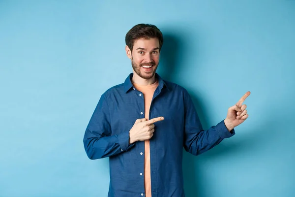 Vrolijke blanke man met baard wijzende vingers opzij naar lege ruimte en glimlachend naar de camera, met reclame, blauwe achtergrond — Stockfoto