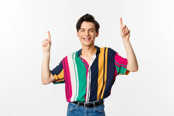Handsome queer man in stylish clothes showing logo, pointing fingers up and smiling pleased, standing over white background — Stock Photo, Image
