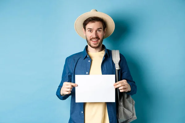 Alegre turista en sombrero de verano, mostrando un pedazo de papel vacío y sonriente, autoestop con mochila, de pie sobre fondo azul — Foto de Stock