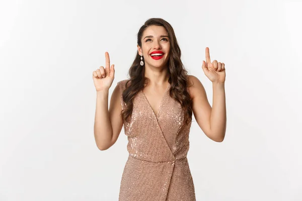 Año Nuevo, Navidad y concepto de celebración. Mujer elegante feliz en vestido de fiesta, riendo y mirando hacia arriba, apuntando a la oferta promocional superior, fondo blanco —  Fotos de Stock