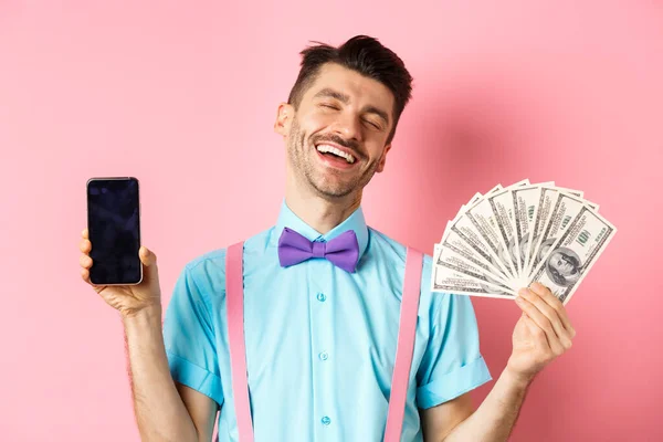 Concepto de comercio electrónico y compras. Rico y feliz chico riendo, mostrando dinero en efectivo y pantalla vacía del teléfono inteligente, de pie sobre fondo rosa — Foto de Stock
