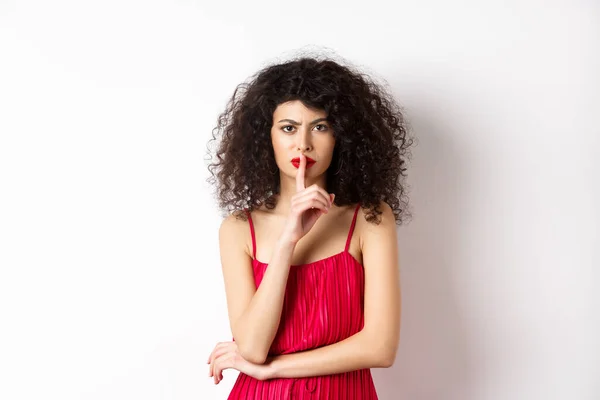 Θυμωμένη κομψή γυναίκα σε κόκκινο φόρεμα hushing και συνοφρύωμα, πείτε να είναι ήσυχο, ζητώντας σιωπή, στέκεται πάνω από το λευκό φόντο — Φωτογραφία Αρχείου