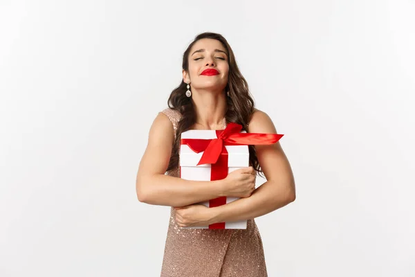 Vacaciones, concepto de celebración. Mujer feliz y agradecida abrazando regalo de Navidad, usando vestido de lujo, recibiendo regalo de Año Nuevo, de pie sobre fondo blanco — Foto de Stock