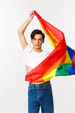 Ekin bluzlu ve kot pantolonlu mutlu eşcinsel kişinin dikey görüntüsü gökkuşağı bayrağını sallıyor, lgbtq bayramını kutluyor, beyaz arka planda duruyor.