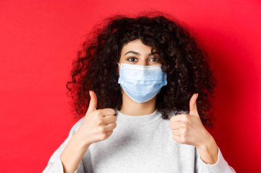 Covid-19 ve pandemik konsept. Yüz maskeli genç, kıvırcık bir kadının yakından çekilmiş fotoğrafı. Kırmızı arka planda duran güzel bir şey.