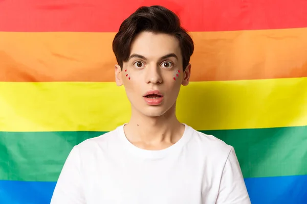 Primer plano de la persona queer sorprendido con brillo en la cara, mirando asombrado a la cámara, de pie sobre la bandera del arco iris lgbtq —  Fotos de Stock