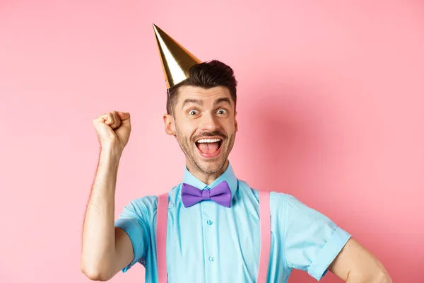 Vakantie en feest concept. Gelukkige verjaardag jongen met snor en strik viering evenement in feestkegel hoed en feestelijke kleding, het opheffen van de hand omhoog en schreeuwen ja — Stockfoto